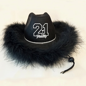 May include: Black cowboy hat with a black feather trim. The hat has a white embroidered design that says "21 Finally".