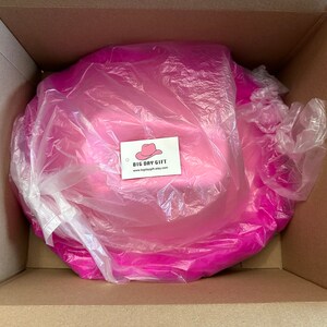 Peut inclure: Un chapeau de cow-boy rose est emball&eacute; dans une bo&icirc;te en carton avec un sac en plastique blanc. Le chapeau est envelopp&eacute; dans du papier de soie rose. La bo&icirc;te porte une &eacute;tiquette indiquant "BIG DAY GIFT" avec une adresse de site Web.