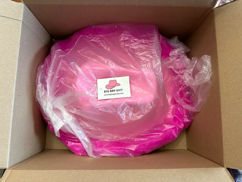 Peut inclure: Un chapeau de cow-boy rose est emball&eacute; dans une bo&icirc;te en carton avec un sac en plastique blanc. Le chapeau est envelopp&eacute; dans du papier de soie rose. La bo&icirc;te porte une &eacute;tiquette indiquant "BIG DAY GIFT" avec une adresse de site Web.