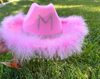 May include: Pink cowboy hat with a large rhinestone letter M on the front. The hat has a pink feather trim and a rhinestone band.