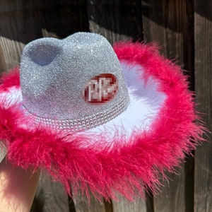 May include: A silver glitter cowboy hat with a white brim and a pink feather trim. The hat has a red and white sticker that says "PPC".