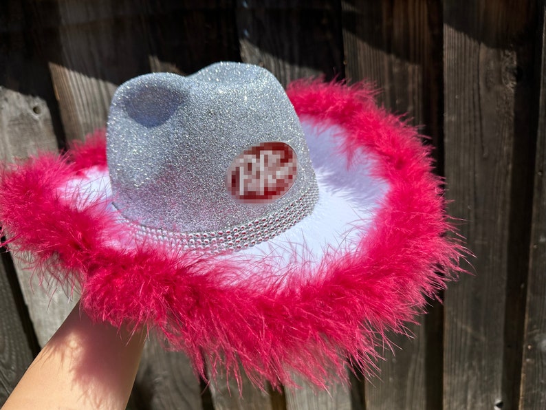 May include: A silver glitter cowboy hat with a white brim and a pink feather trim. The hat has a red and white sticker that says "PPC".