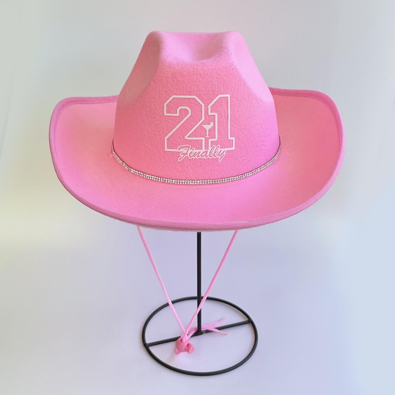 Peut inclure: Chapeau de cow-boy rose avec une bande de strass blanche et le chiffre 21 avec le mot "Finally" en dessous.