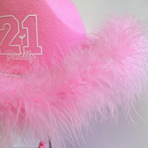 Peut inclure: Chapeau de cow-boy rose avec une garniture de plumes blanches et le num&eacute;ro 21 avec le mot "Enfin" &eacute;crit en dessous.