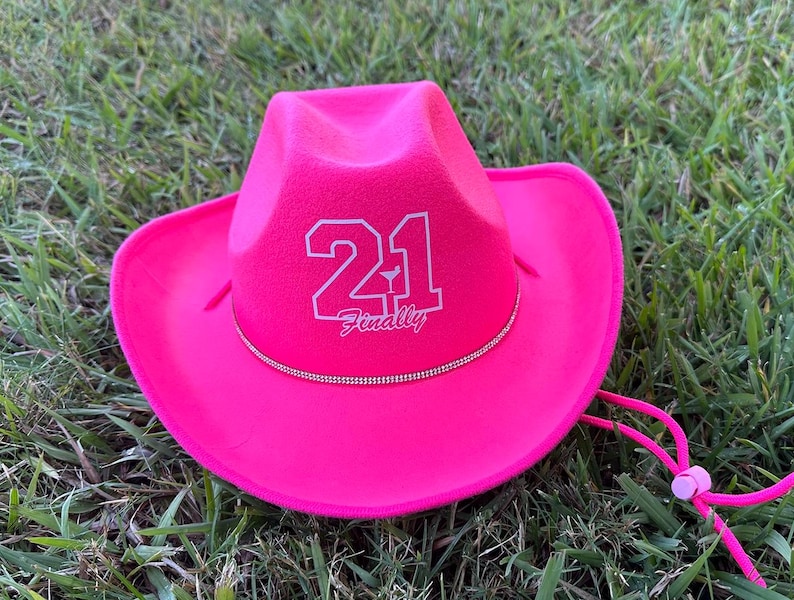 Peut inclure: Un chapeau de cowboy rose vif avec un design en feutre blanc qui dit "21 Enfin" avec un petit graphique de verre &agrave; cocktail.