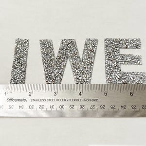 May include: Silver glitter letters spelling out "IWE" on a white background. The letters are approximately 2 inches tall and are measured with a ruler.