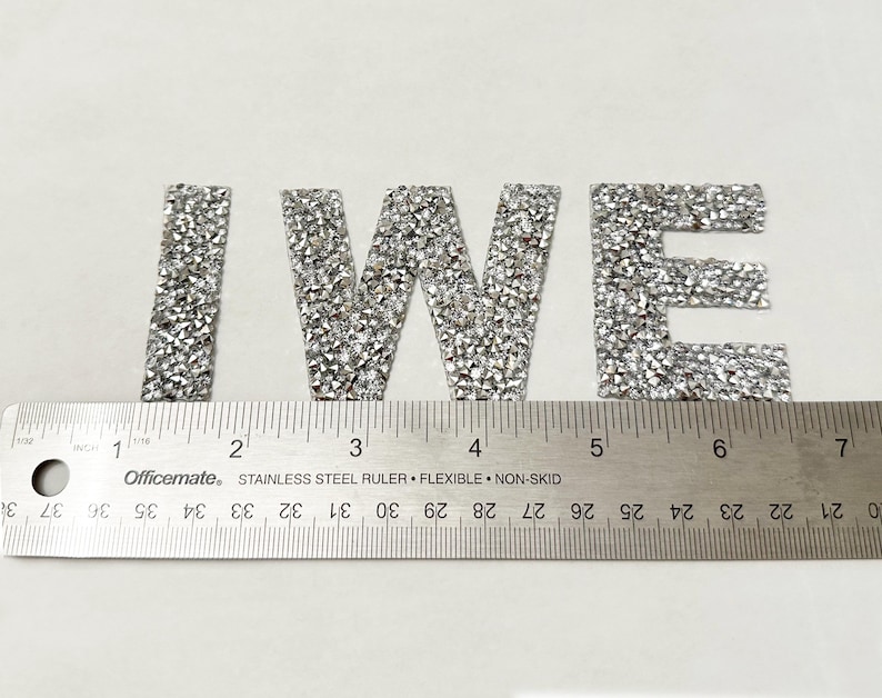 May include: Silver glitter letters spelling out "IWE" on a white background. The letters are approximately 2 inches tall and are measured with a ruler.