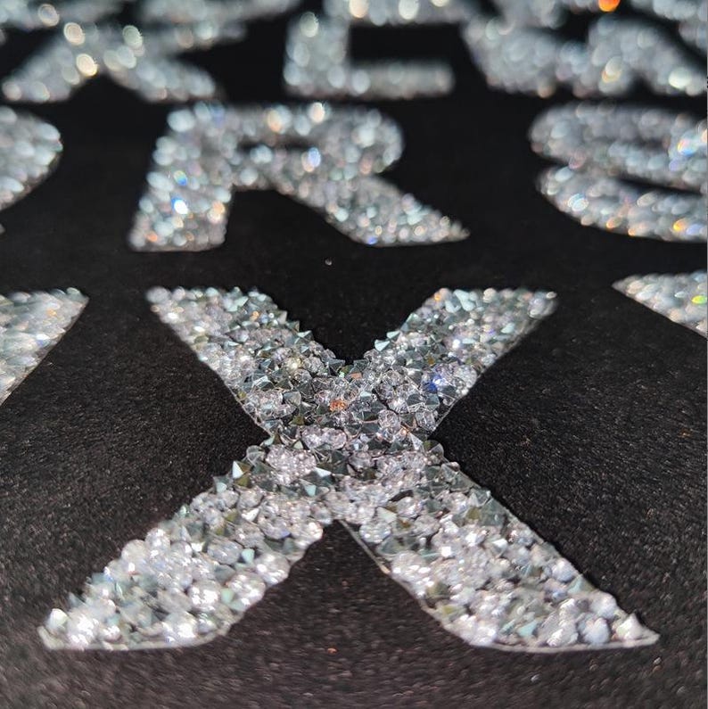May include: A close-up of a black fabric with the letter "X" made of small, clear rhinestones.