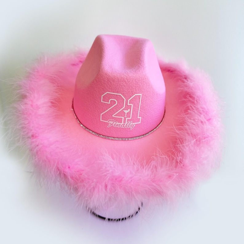 Peut inclure: Chapeau de cow-boy rose avec une bordure en plumes blanches et le num&eacute;ro 21 avec le mot "finally" &eacute;crit en dessous.