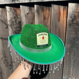 May include: A green cowboy hat with a Jameson Irish Whiskey label on the front. The hat is decorated with silver glitter and has a fringe of silver beads around the brim.