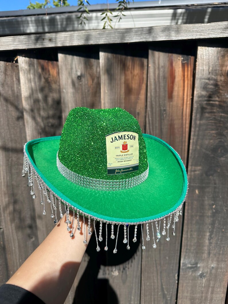 May include: A green cowboy hat with a Jameson Irish Whiskey label on the front. The hat is decorated with silver glitter and has a fringe of silver beads around the brim.