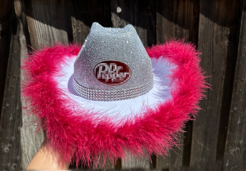 May include: A silver glitter cowboy hat with a white band and pink feather trim. The hat has a red and white oval logo that says "Dixie" on it.