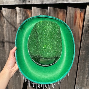 May include: A green felt hat with a green glitter top and a silver trim. The hat has a silver label that says "Jameson" and a fringe of silver beads around the bottom.