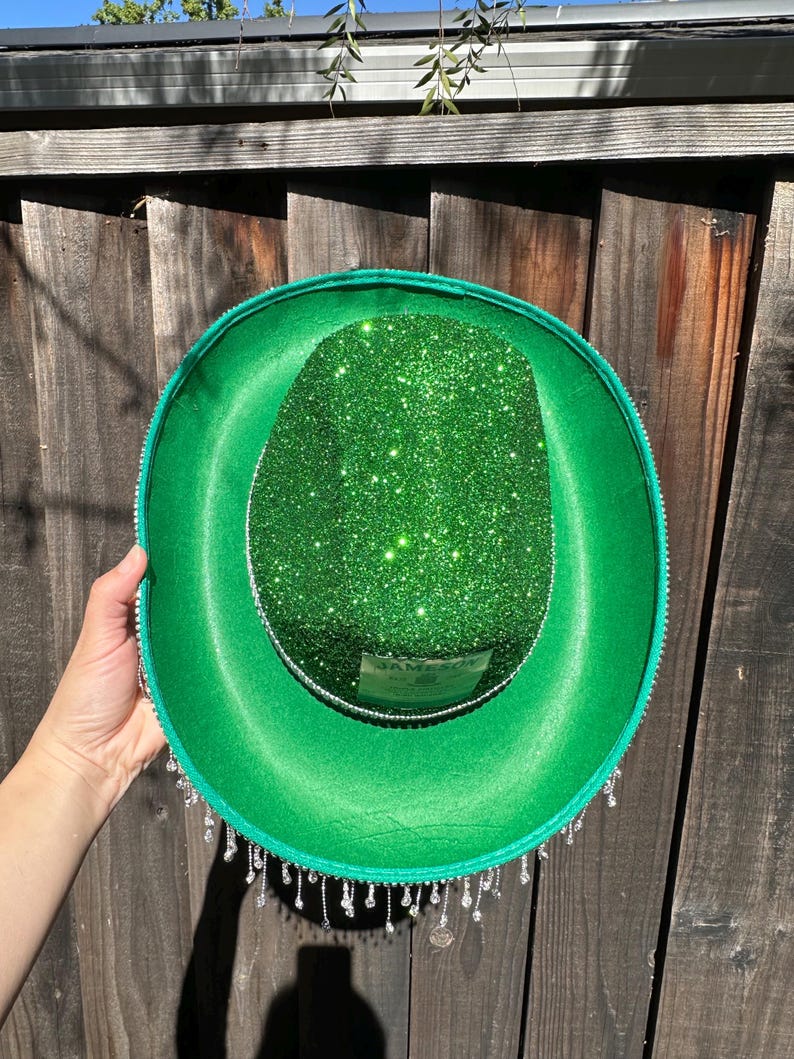 May include: A green felt hat with a green glitter top and a silver trim. The hat has a silver label that says "Jameson" and a fringe of silver beads around the bottom.