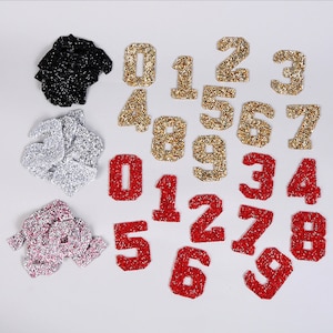 May include: Assortment of iron-on glitter numbers in red, gold, and silver. The numbers range from 0 to 9.