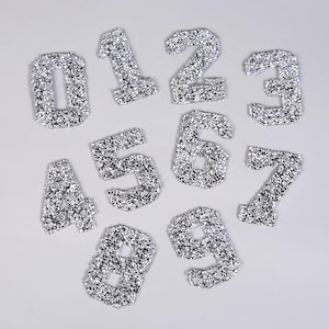 May include: Ten silver glitter iron-on patches in the shape of numbers 0 through 9.
