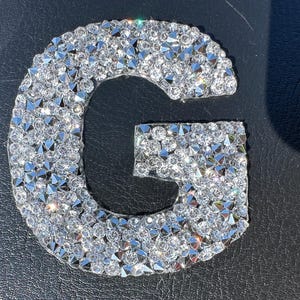 May include: A silver letter G made of rhinestones. The letter is on a black background.