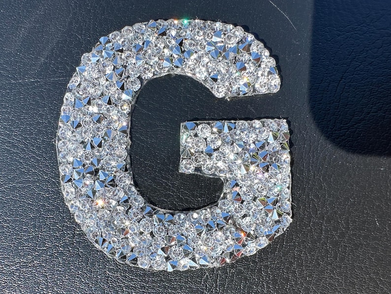 May include: A silver letter G made of rhinestones. The letter is on a black background.