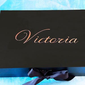 Puede incluir: Una caja de regalo negra con una cinta y un lazo azul marino. La caja tiene el nombre "Victoria" escrito en letras doradas en la parte superior.
