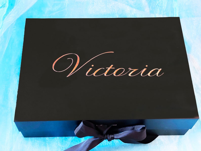 Puede incluir: Una caja de regalo negra con una cinta y un lazo azul marino. La caja tiene el nombre "Victoria" escrito en letras doradas en la parte superior.