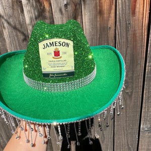 May include: Green cowboy hat with a silver glitter crown and a silver fringe. The hat has a Jameson Irish Whiskey label on the front.
