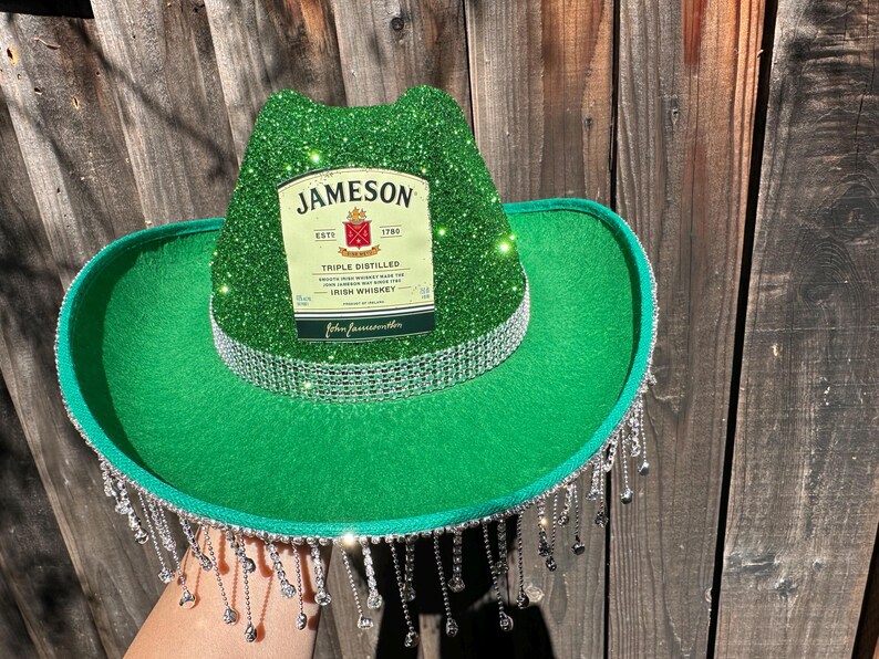 May include: Green cowboy hat with a silver glitter crown and a silver fringe. The hat has a Jameson Irish Whiskey label on the front.