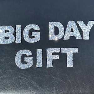 May include: A black background with the words "BIG DAY GIFT" in silver glitter letters.