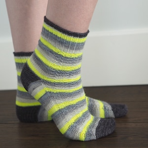 Könnte beinhalten: Ein Paar gestreifte Socken in den Farben Grau, Schwarz und Neon-Gelb. Die Socken sind aus weichem, gestricktem Stoff und haben eine bequeme Passform.