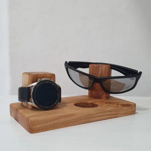 May include: A wooden organizer with a watch and sunglasses. The watch has a black band and a silver face. The sunglasses are black. The organizer is made of light-colored wood and has a circular hole.
