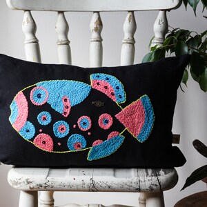 May include: Black rectangular pillow with a colorful embroidered fish design. The fish features blue, pink, and yellow stitching on a black background. The pillow is placed on a white wooden chair.