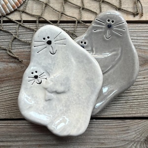 May include: Two ceramic seal-shaped dishes with a light brown and a gray glaze. The dishes are on a wooden surface with seashells and a starfish.