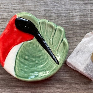 May include: A ceramic dish shaped like a hummingbird with red, white, and green glaze. The dish is sitting on a wooden surface next to a tea bag with a gold tag that says "Over 300 Years of Experience est. 1706".