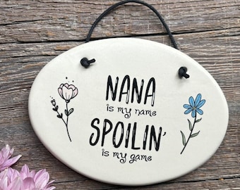 May include: White oval ceramic sign with black text that reads "NANA is my name SPOILIN' is my game". The sign has a black string for hanging and features a pink flower on the left and a blue flower on the right.