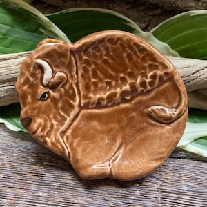 May include: A brown ceramic bison sculpture with a white horn. The bison is lying down with its head turned to the side. The sculpture is detailed with textured fur and a realistic face.