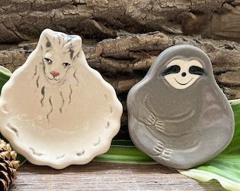 Può includere: Due piattini in ceramica per piccoli oggetti, uno a forma di bradipo con un viso sorridente e l'altro a forma di lama con il naso rosa. Entrambi i piattini sono dipinti a mano e hanno un aspetto rustico e artigianale.