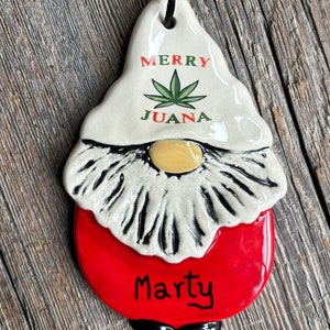 May include: A white ceramic gnome ornament with a red hat and a green marijuana leaf. The ornament says "Merry Juana" and "Marty" on the front.