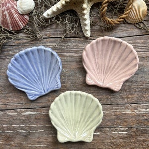 May include: Three ceramic seashell-shaped dishes in different colors: light blue, light pink, and cream. The dishes are arranged on a wooden surface.