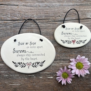 May include: Two white oval ceramic signs with black hanging cords. The signs have a black floral design and a pink heart. The text on the signs reads "Side by Side or miles apart. Sisters always stay connected by the heart."