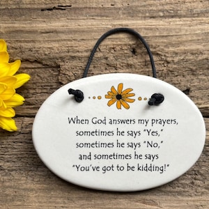 May include: White ceramic oval ornament with a black string for hanging. It features a yellow flower and the text "When God answers my prayers, sometimes he says "Yes," sometimes he says "No," and sometimes he says "You've got to be kidding!"