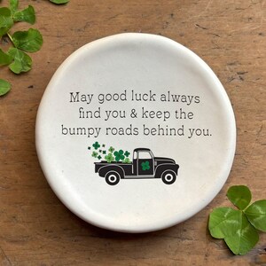 May include: White ceramic dish with a black truck carrying green four-leaf clovers. The dish has the text "May good luck always find you & keep the bumpy roads behind you."