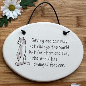 May include: White ceramic oval sign with a black string for hanging. The sign has a black line drawing of a cat sitting and the text "Saving one cat may not change the world but for that one cat, the world has changed forever."