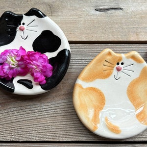 May include: Two ceramic cat-shaped dishes, one black and white, the other orange and white, with painted details and a happy face.