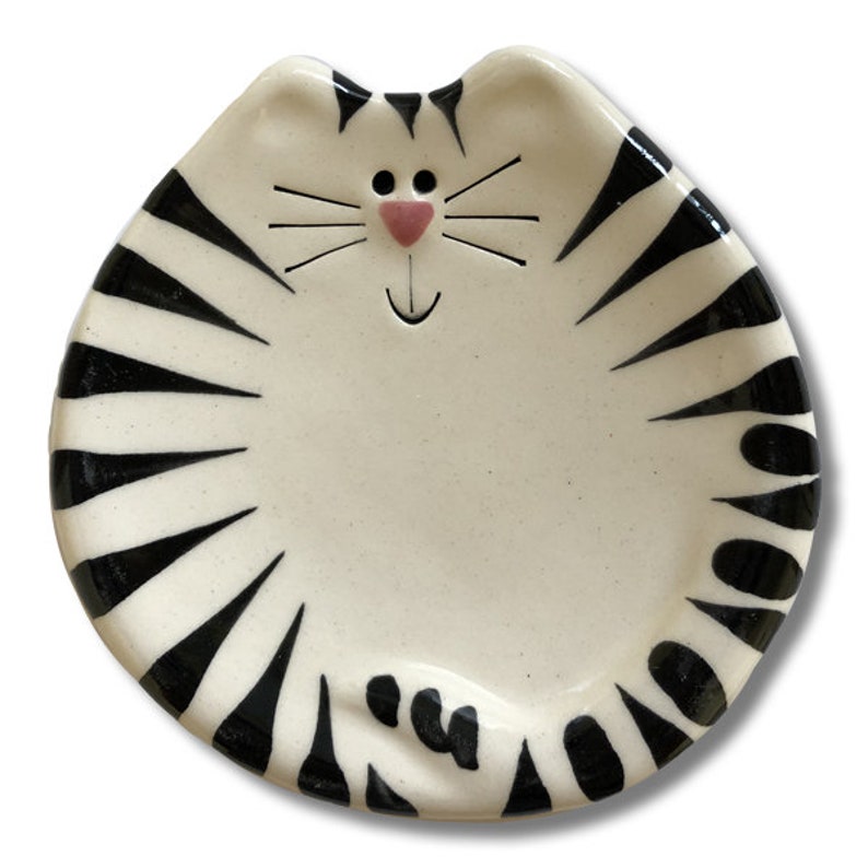 May include: A ceramic dish shaped like a cat with black and white stripes. The cat has a pink nose and a smiling mouth.