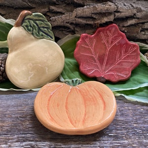 May include: Three ceramic dish trivets shaped like a pear, a leaf, and a pumpkin. The pear is yellow with a green stem and leaf. The leaf is red with a detailed vein pattern. The pumpkin is orange with a green stem.