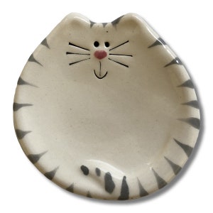 May include: A white ceramic dish shaped like a cat with gray stripes. The cat has a pink nose and a black smiling mouth.