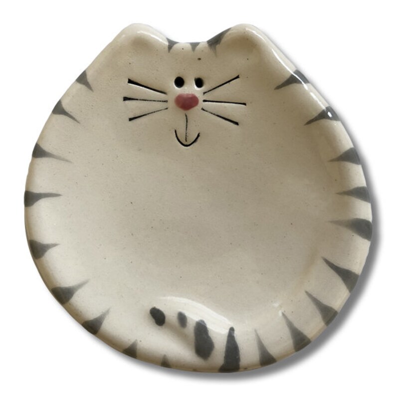 May include: A white ceramic dish shaped like a cat with gray stripes. The cat has a pink nose and a black smiling mouth.