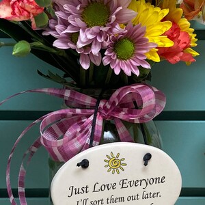 May include: A glass jar with a pink and white striped ribbon tied around the neck. The jar holds a bouquet of yellow, pink, and red flowers. A white oval ceramic tag with a yellow sun and the text "Just Love Everyone I'll sort them out later. -God" is attached to the jar.