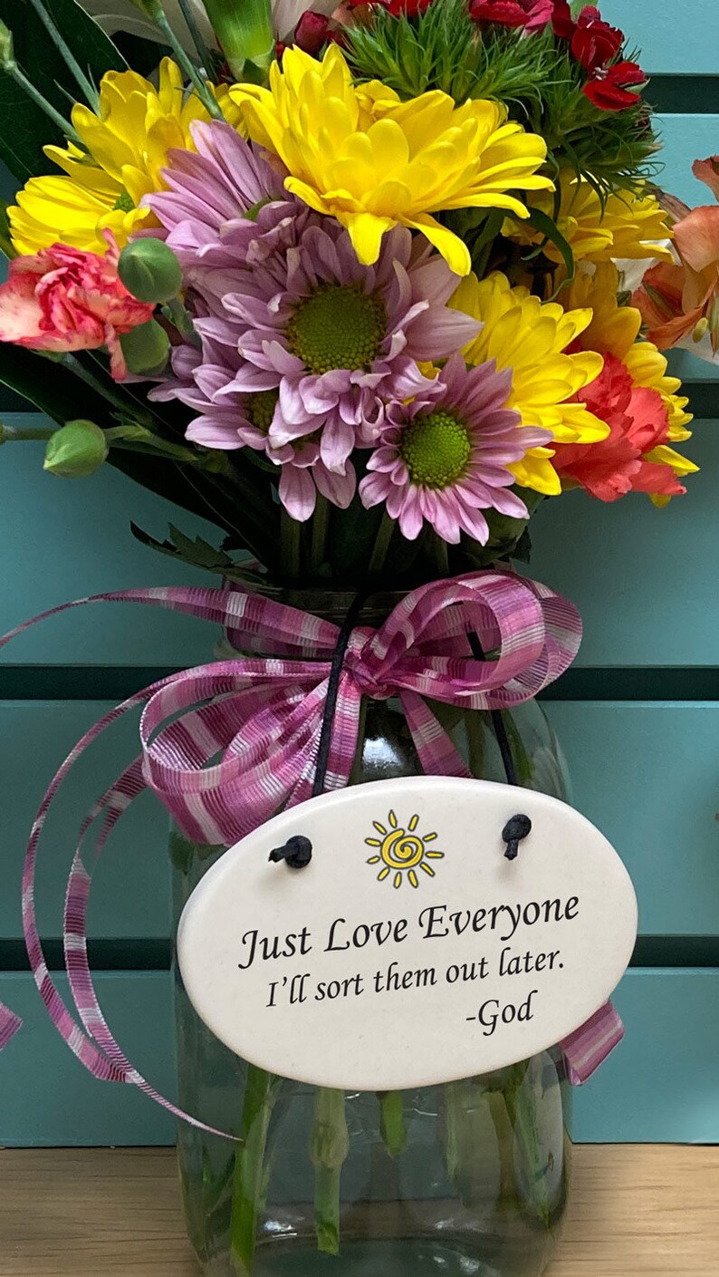May include: A glass jar with a pink and white striped ribbon tied around the neck. The jar holds a bouquet of yellow, pink, and red flowers. A white oval ceramic tag with a yellow sun and the text "Just Love Everyone I'll sort them out later. -God" is attached to the jar.