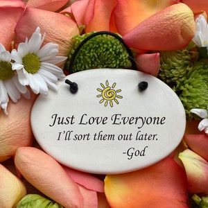 May include: White ceramic oval ornament with a black string for hanging. The ornament has a yellow sun with a spiral design and the text "Just Love Everyone I'll sort them out later. -God". The ornament is surrounded by pink and white flower petals.