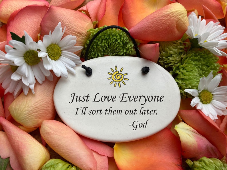 May include: White ceramic oval ornament with a black string for hanging. The ornament has a yellow sun with a spiral design and the text "Just Love Everyone I'll sort them out later. -God". The ornament is surrounded by pink and white flower petals.